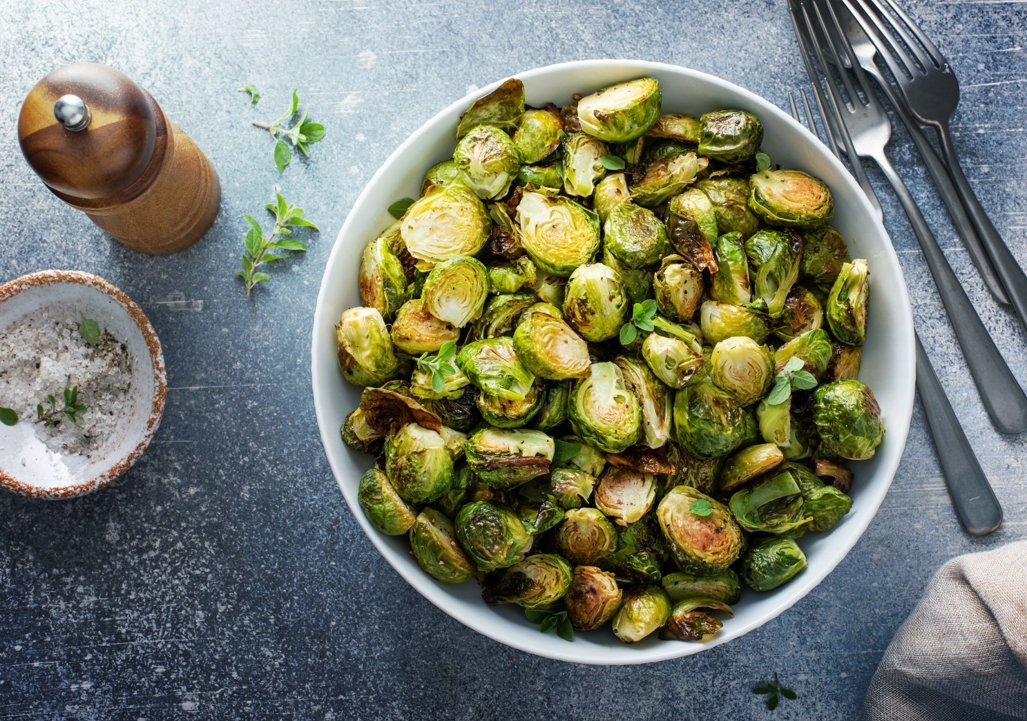 Roasted Brussels sprouts in a bowl on a table with forks, bowl of salt and pepper mill